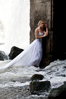 Young blonde woman in a white wedding dress near the waterfall