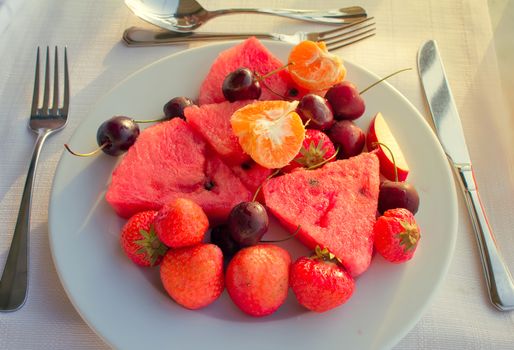 Strawberries, cherries, slices of watermelon, slices of Mandarin are located on a plate on a white tablecloth table.