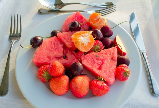 Strawberries, cherries, slices of watermelon, slices of Mandarin are located on a plate on a white tablecloth table.