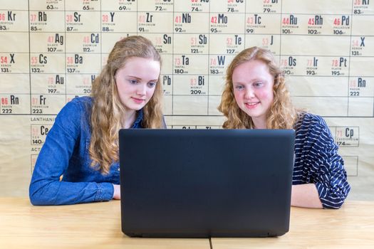 Two caucasian teenage girls looking at laptop in chemistry class in front of wallchart showing periodic table