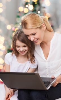 family, childhood, holidays, technology and people - smiling mother and little girl with laptop computer over christmas tree lights background