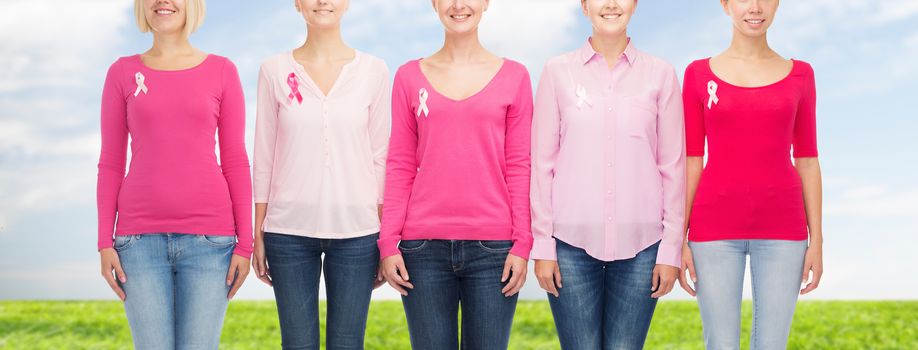 healthcare, people and medicine concept - close up of smiling women in blank shirts with pink breast cancer awareness ribbons over blue sky and grass background