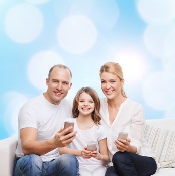 family, holidays, technology and people concept - smiling mother, father and little girl with smartphones over blue lights background