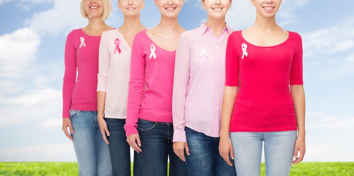 healthcare, people and medicine concept - close up of smiling women in blank shirts with pink breast cancer awareness ribbons over blue sky and grass background