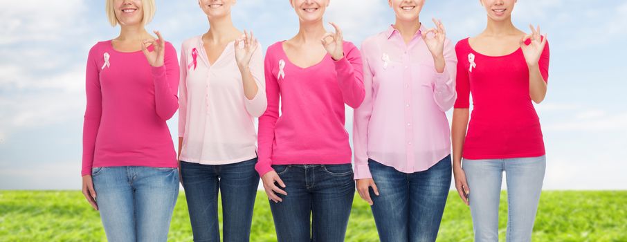 healthcare, people, gesture and medicine concept - close up of smiling women in blank shirts with pink breast cancer awareness ribbons showing ok sign over blue sky and grass background