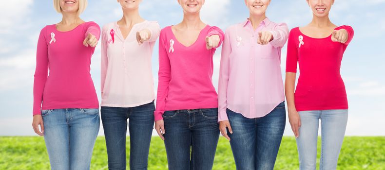 healthcare, people, gesture and medicine concept - close up of smiling women in blank shirts with pink breast cancer awareness ribbons pointing on you over blue sky and grass background