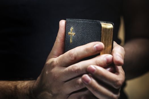 Man holding an old small black bible in his hands. Short depth of field, the sharpness is in the cross.
