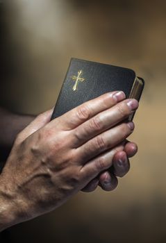 Man holding an old small black bible in his hands. Short depth of field, the sharpness is in the cross.