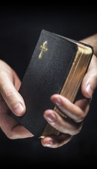 Man holding an old small black bible in his hands. Short depth of field, the sharpness is in the cross.