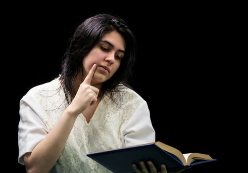 Latina teen in white dress holding a blue book with gold edging, holding a finger to her cheek and standing in front of a black background