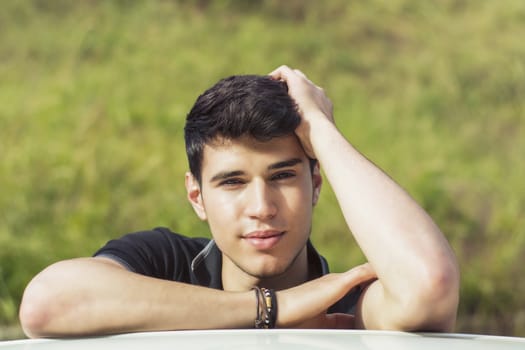 Head and arms shot of handsome attractive young man looking at camera outdoor, leaning on flat surface with head resting on hands