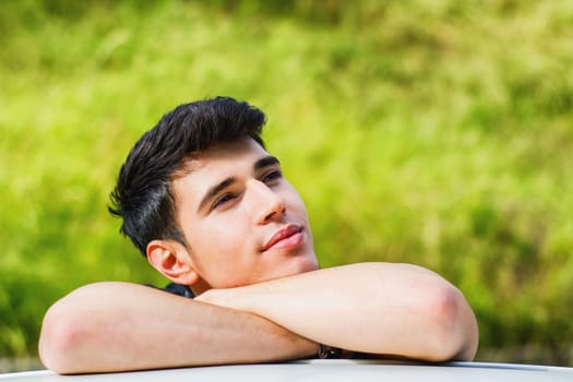 Head and arms shot of handsome attractive young man looking at camera outdoor, leaning on flat surface with head resting on hands