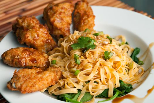 Thai style fried chicken wings on a round white plate with egg noodles and spinach. Shallow depth of field.