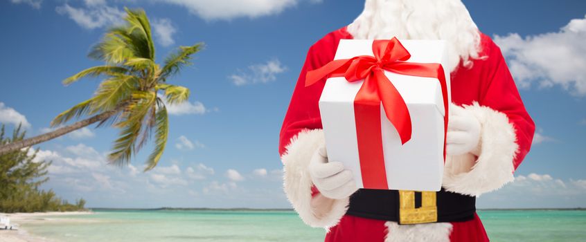 christmas, holidays, travel and people concept - close up of santa claus with gift box over tropical beach background