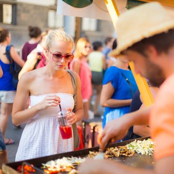 Beautiful blonde caucasian lady wearing white summer dress buying a meal freshly prepared at a local food market. Urban international kitchen event taking place in Ljubljana, Slovenia, in summertime.