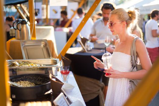 Beautiful blonde caucasian lady wearing white summer dress buying freshly prepared mealat a local food market. Urban international kitchen event taking place in Ljubljana, Slovenia, in summertime.