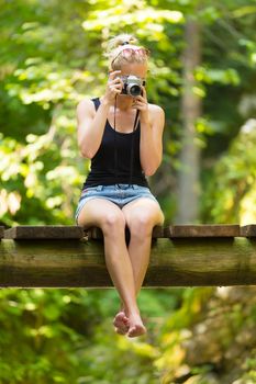 Beautiful barefooted blonde caucasian girl wearing jeans shorts an sporty black sleeveless t-shirt, sitting on a vintage wooden bridge, taking photos with retro camera.