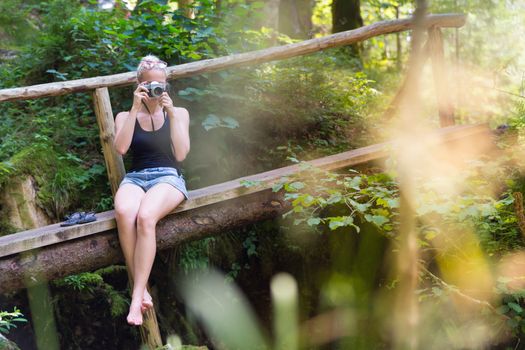 Beautiful barefooted blonde caucasian girl wearing jeans shorts an sporty black sleeveless t-shirt, sitting on a vintage wooden bridge, taking photos with retro camera.