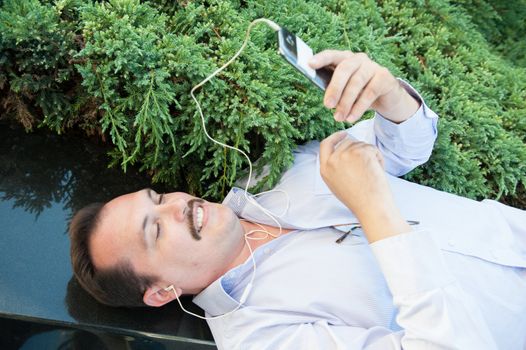 Urban man using smart phone outside using app on 4g wireless device wearing headphones. Lying on the Marble fence