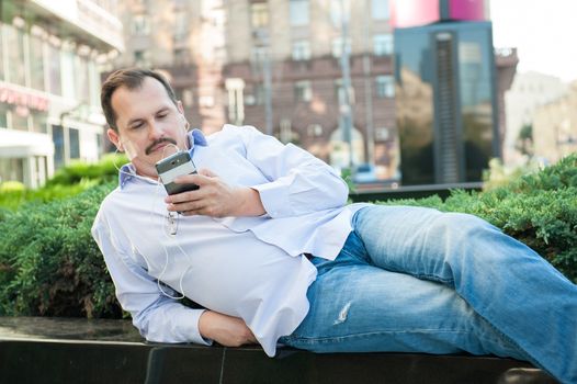 Urban man using smart phone outside using app on 4g wireless device wearing headphones. Lying on the Marble fence