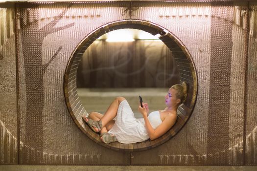 Lady wearing white summer dress playing carelessly with her android smart phone browsing trough social networks relaxing on contemporary circular bench in urban pedestrian underpass. 