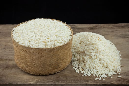 Rice in a wicker rice and scattered near on dark wooden table.