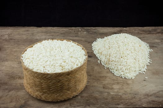 Rice in a wicker rice and scattered near on dark wooden table.