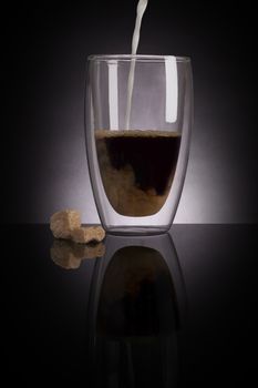 Pouring milk into filter coffee in glass on dark black background. Delicious coffee drinking.