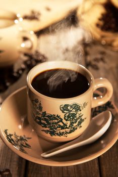 Steaming traditional Chinese nanyang style coffee in vintage mug and saucer with coffee beans. Fractal on the cup is generic print. Soft focus setting with dramatic ambient light on dark wooden background.