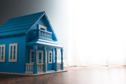 Blue wooden model house next to a window with curtain on wooden floor.