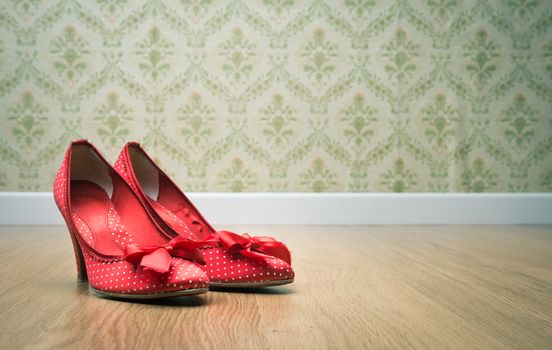 Vintage red dotted female shoes with ribbon on hardwood floor with retro wallpaper on background.