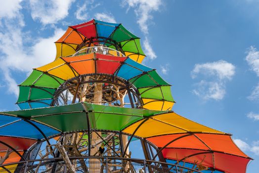 A tall, bright and colorful jungle gym built around some tree trunks on a blue sky background dotted with clouds