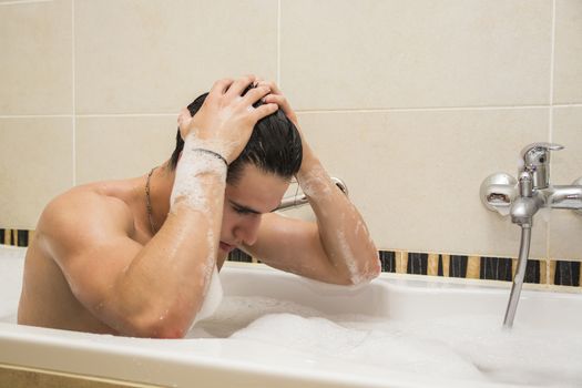 Handsome young man in bathtub at home having bath, washing body and hair with bathfoam and shampoo