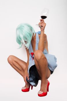 Attractive model woman in blue wig and red high heels posing sitting with glass of wine isolated on white