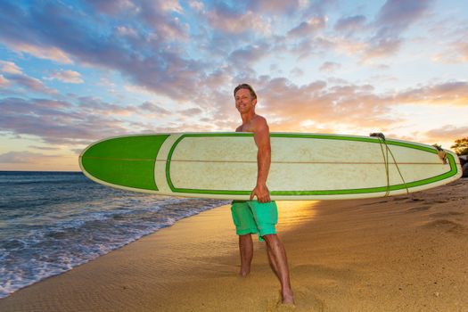 Athletic upbeat adult holding his surfboard on beach at sunset