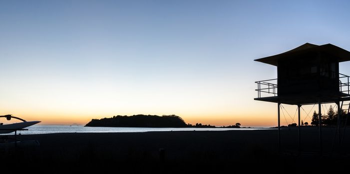 Silhouetted against golden glow of sunrise lifesafers lookout on Mount Maunganui beach New Zealand.