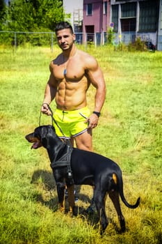 Muscular shirtless man with dobermann dog at leash, walking in city park