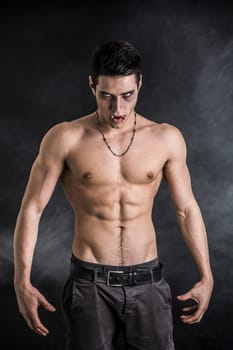 Portrait of a Young Vampire Man Shirtless, Showing his Torso, Chest and Abs, Looking at the Camera, on Dark Background.