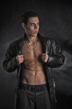 Portrait of a Young Vampire Man in an Open Black Leather Jacket, Showing his Chest and Abs, Looking to Right, on Dark Background.