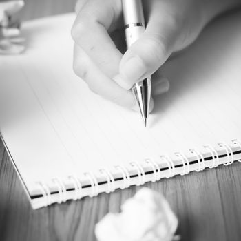 woman hand writing with pen on notebook.there are crumpled paper and coffee cup on wood table background black and white color tone style