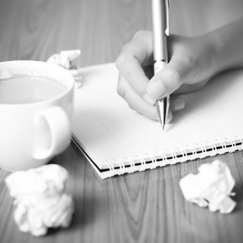 woman hand writing with pen on notebook.there are crumpled paper and coffee cup on wood table background black and white color tone style