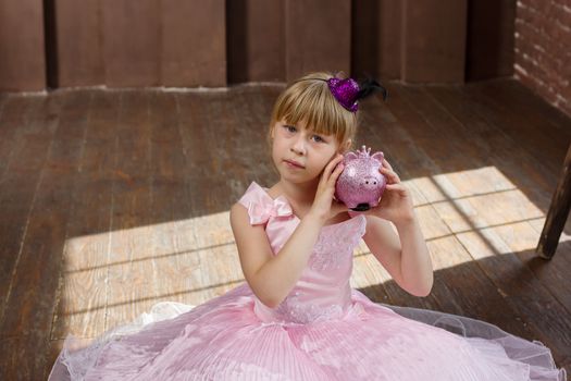 Girl 6 years old in a pink dress with a piggy bank in his hands. In Room. Horizontal framing