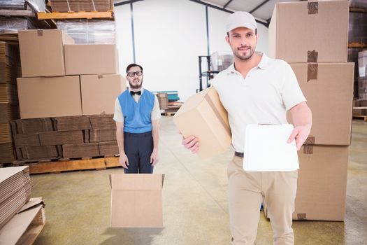 Delivery man with cardboard box showing clipboard  against boxes on trolley in warehouse