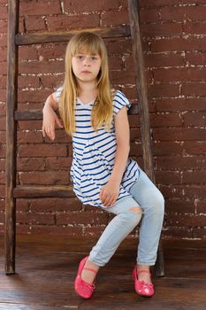 Girl 6 years old in jeans sits on a ladder near a brick wall