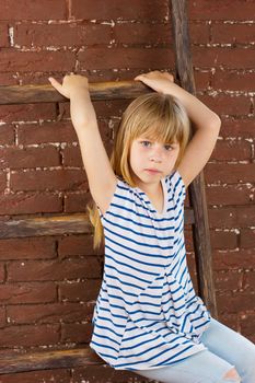 Girl 6 years old in jeans and a vest sits on a ladder near a brick wall. Vertical framing