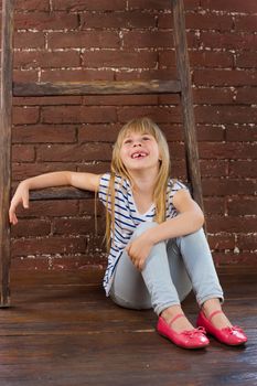 Girl 6 years old in jeans and a vest sits on the floor next to a brick wall and filled with laughter. Vertical framing