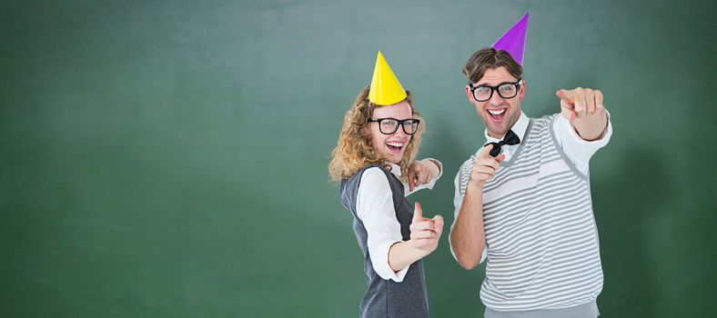 Happy geeky hispser couple dancing with party hat against green chalkboard