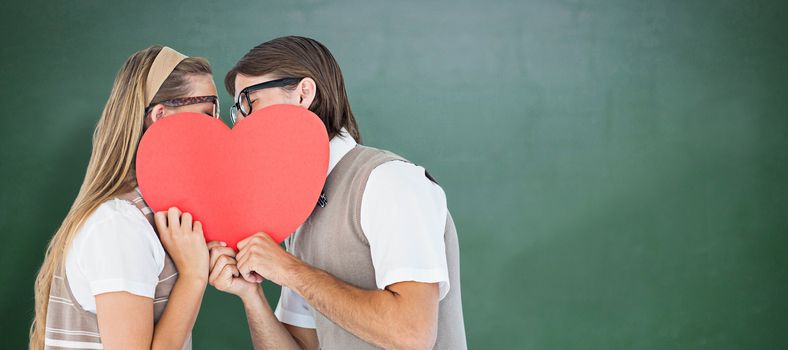 Geeky hipsters kissing behind heart card  against green chalkboard
