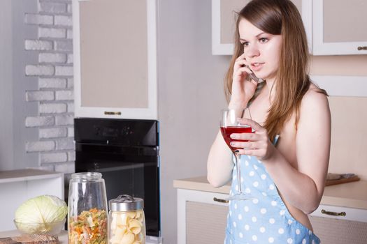 girl in the kitchen wearing an apron over his naked body with a glass of wine on the phone