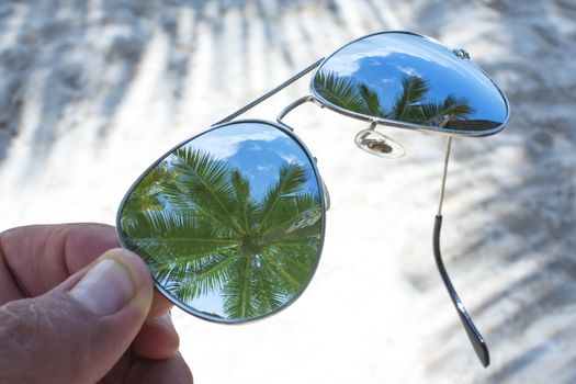 Closeup on lens of reflective sunglasses over sandy beach reveals reflections of tall coconut palms in tropical paradise